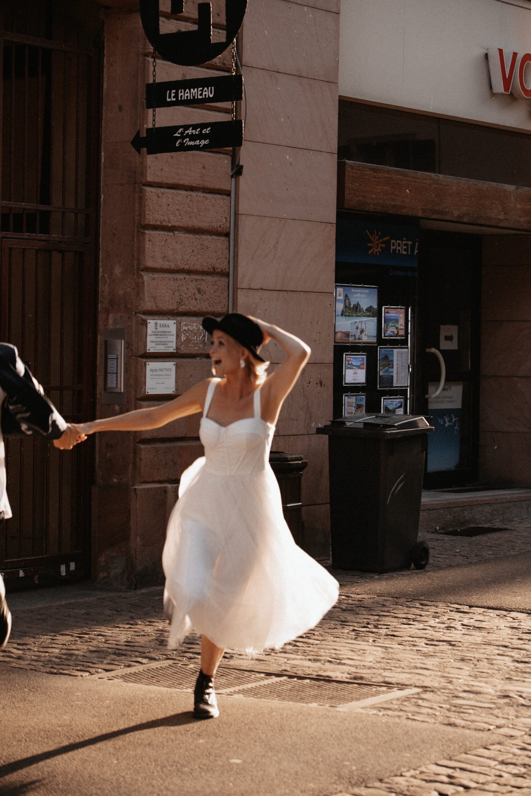 Künstlerpaar bei ihrem Brautpaarshooting in Colmar während es sich küsst, durch die Straßen läuft und rennt und lacht. Sie trägt ein weißes kürzeres Kleid, einen schwarzen Hut, schwarze Stiefel, glitzernde Ohrringe und er trägt ganz legere schwarze Vans, weißes Shirt und einen dunkelblauen Anzug.