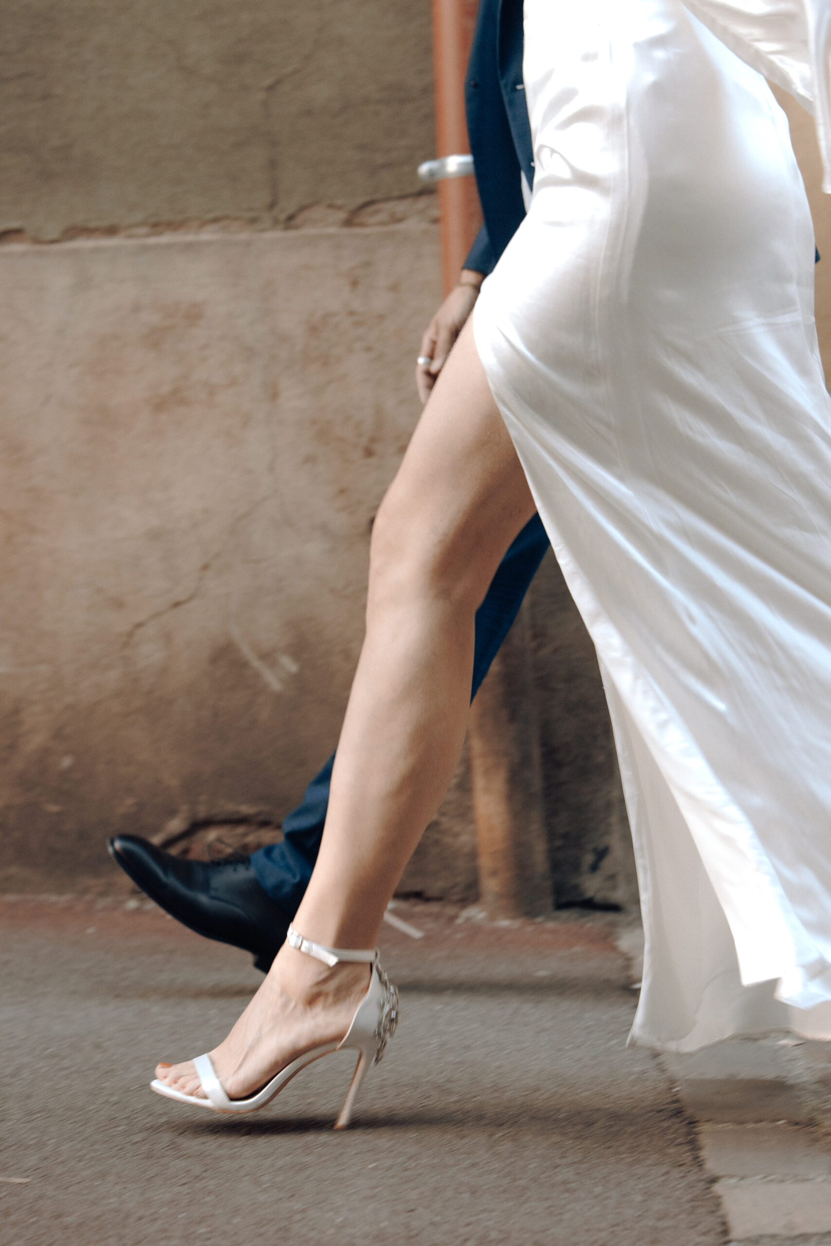 Detailaufnahme eines Braautpaares von den Beinen während sie am Laufen sind. Sie trägt ein weißes seidenes Kleid mit hohen High Heels und das Kleid hat einen Beinschlitz, sodass es gut zum Vorschein kommt.