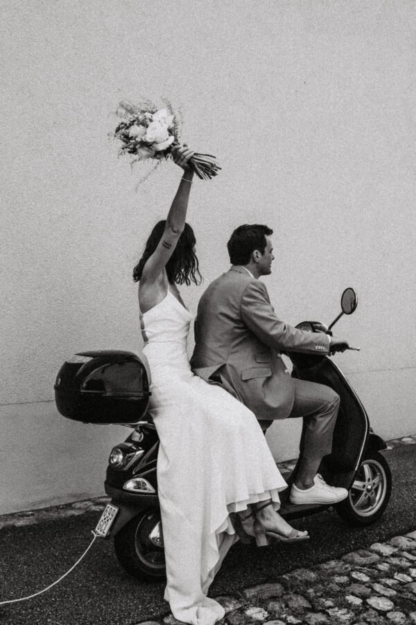schwarz weiß Brautpaarfoto von einem Ehepaar das auf der Vespa vom Standesamt nach Hause fährt und die Braut dabei den Brautstrauß nach oben hält
