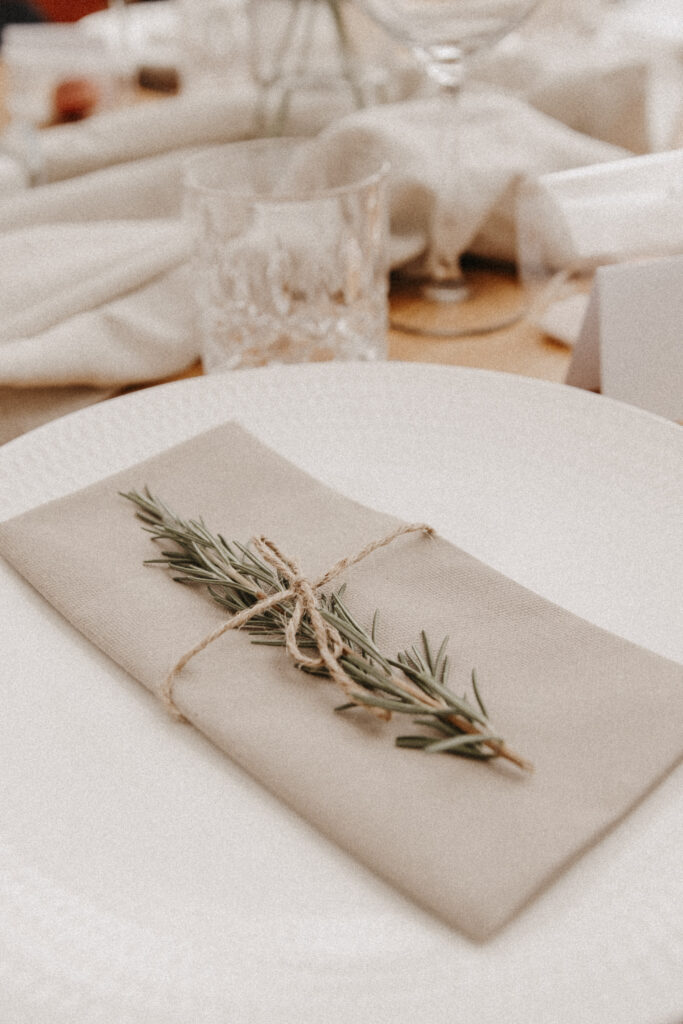 Der Hochzeitsfotograf hat die Hochzeitstischdekoration aufgenommen. Ein weißer Teller mit einer grauen Serviette und einem Rosmarinast ist zu sehen.