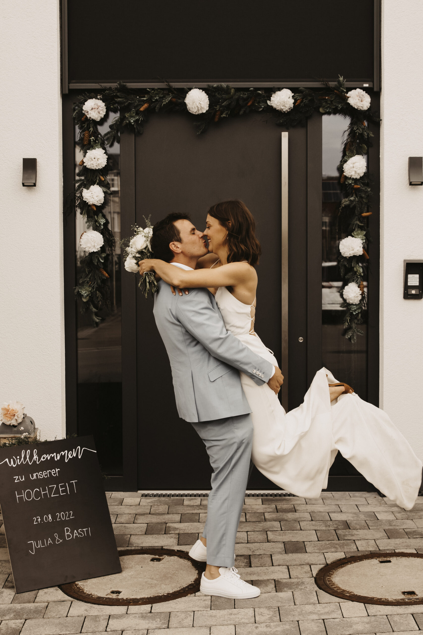 Bräutigam hebt seine Braut hoch und küsst sie dabei vor der dekorierten Hochzeitseingangstüre