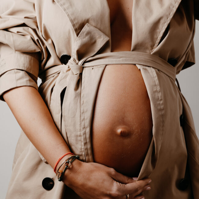 der Fotografie Service Maternityshooting zeigt auf dem Bild eine schwangere Frau von der nur der Bauch zu sehen ist. sie ist umhüllt mit einem beigen Mantel und hält diesen unten zu und der bauch schaut raus.