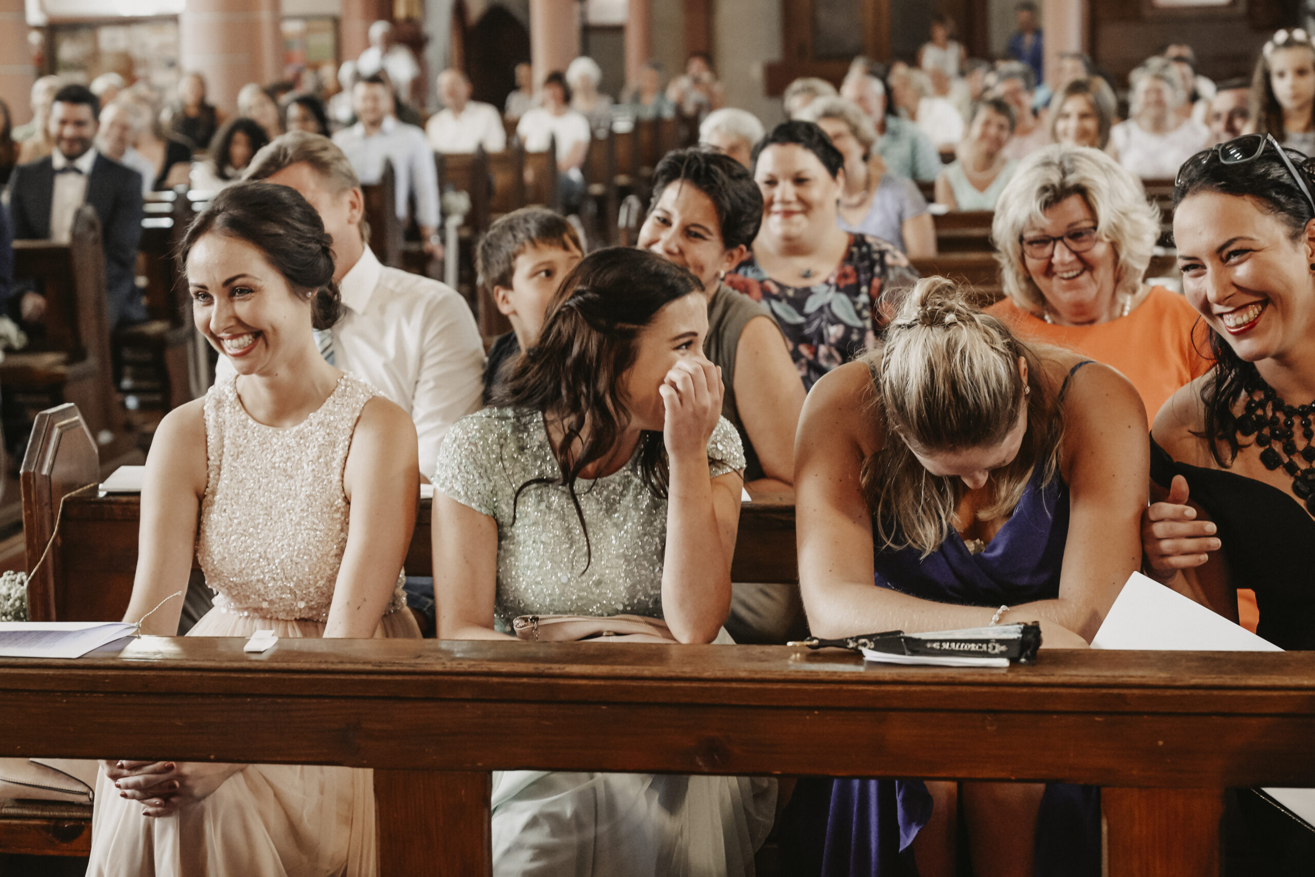 Hochzeitsgäste lachen sich in der Kirche kaputt in der ersten Reihe. Vier Frauen in schönen Kleidern die lachen.