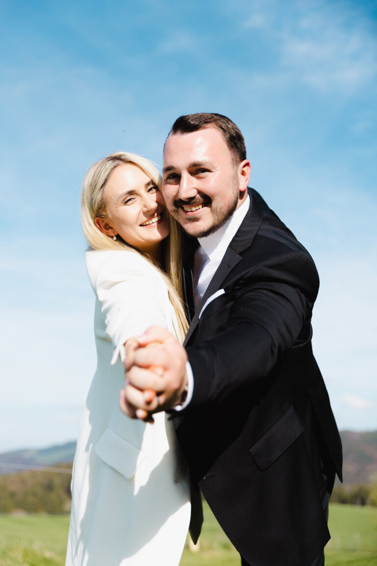 Brautpaar hat Spaß während dem Fotoshooting und macht eine Art Fliegerpose um in die Kamera zu schauen und zu lachen. Die Hochzeitsfotografin spielt mit Schärfe und Unschärfe um das Bild interessanter zu machen. Bild aufgenommen auf dem Schwarzwald - Schauinsland - Haldenköpfle in Farbe.