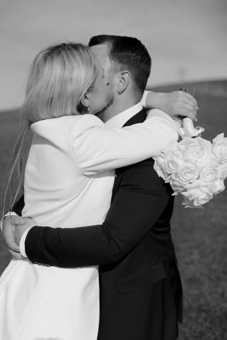Brautpaar küsst und umarmt sich und dabei liegt der Brautstrauß mit den weißen Rosen über seinen Schultern. Bild während einem Brautpaarshooting auf dem Schauinsland. Bild in SW aufgenommen.