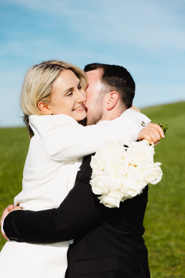 Brautpaar küsst und umarmt sich und dabei liegt der Brautstrauß mit den weißen Rosen über seinen Schultern. Bild während einem Brautpaarshooting auf dem Schauinsland. Bild in Farbe aufgenommen.