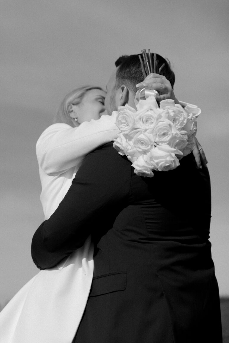 Brautpaar küsst und umarmt sich und dabei liegt der Brautstrauß mit den weißen Rosen über seinen Schultern. Bild in Schwarz weiß aufgenommen während einem Brautpaarshooting auf dem Schauinsland