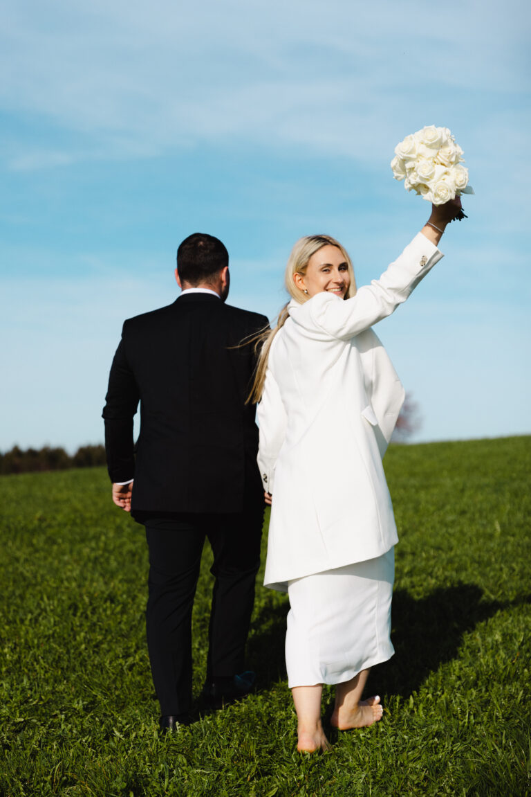 Brautpaar läuft während dem brautpaarshooting auf einer grünen Wiese mit einem blauen Himmel Hintergrund und die Frau hebt den Brautstrauss in die Höhe und schaut dabei über ihre Schulter in die Kamera und lacht
