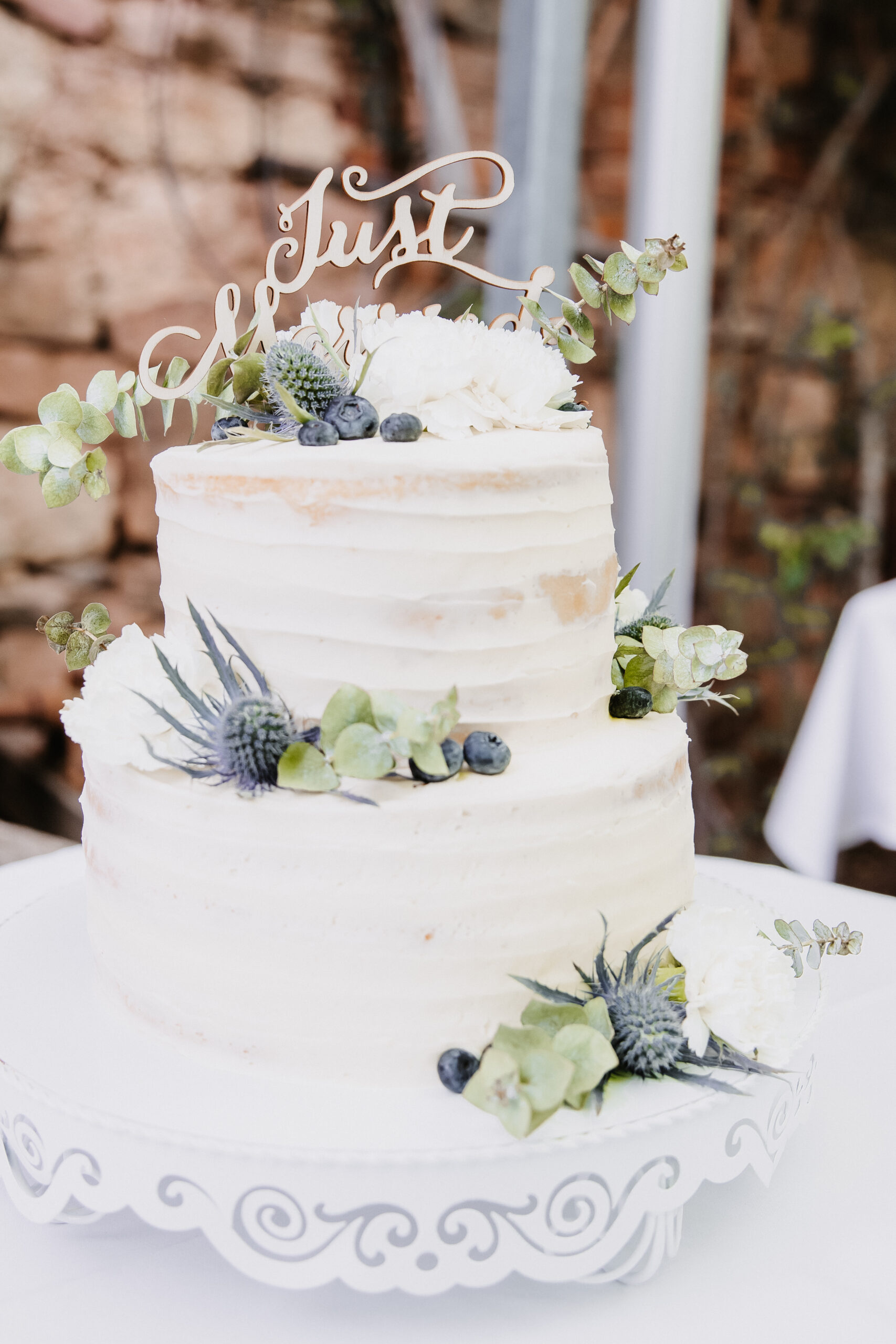 Detailaufnahmen der Hochzeitstorte welche zweiteilig ist mit Heidelbeeren und Trockenblumen sowie einem Just Married Schild.