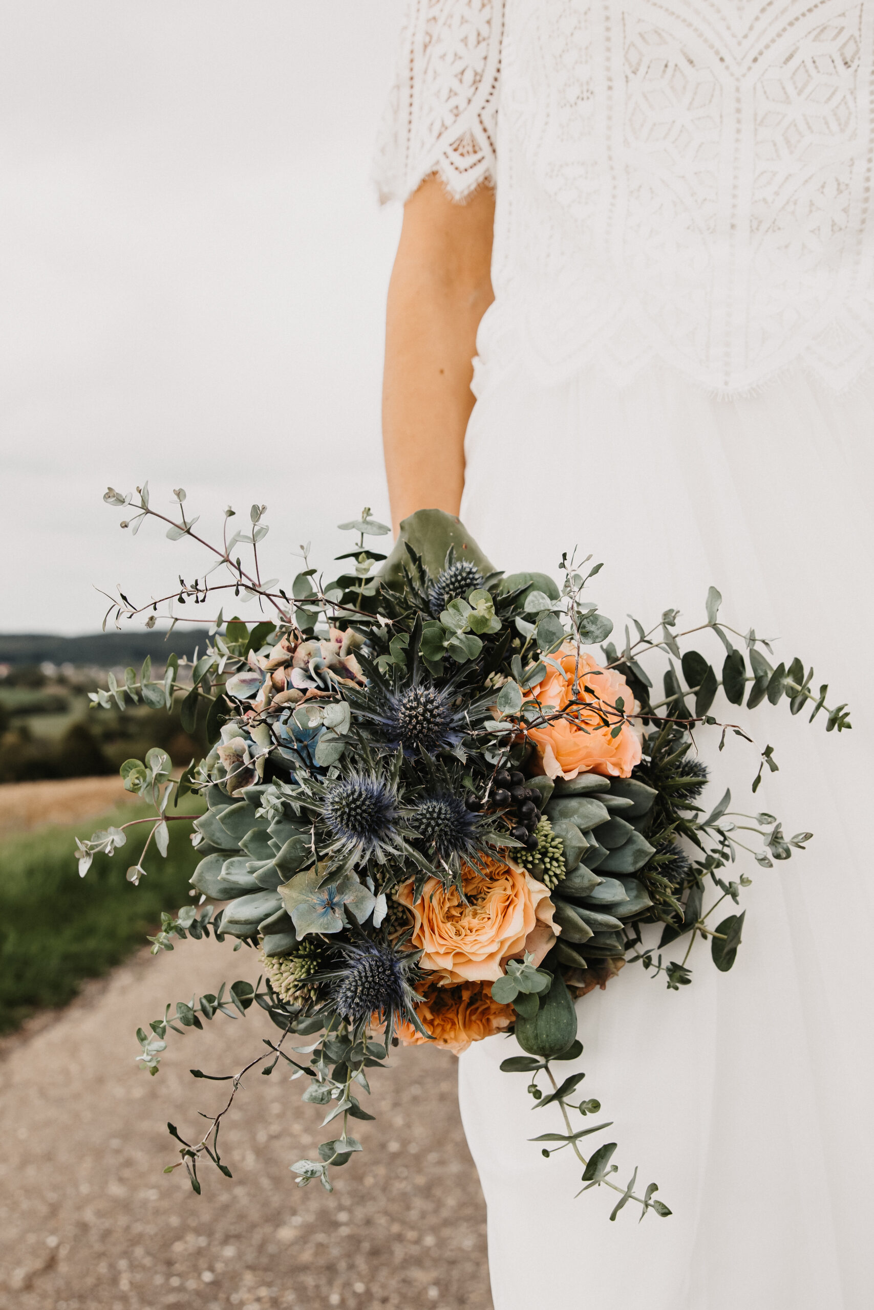 Detailaufnahme des Brautstraußes in Blautönen gehalten mit Aprikot Rosen und die Braut hält ihn an ihrem Körper.