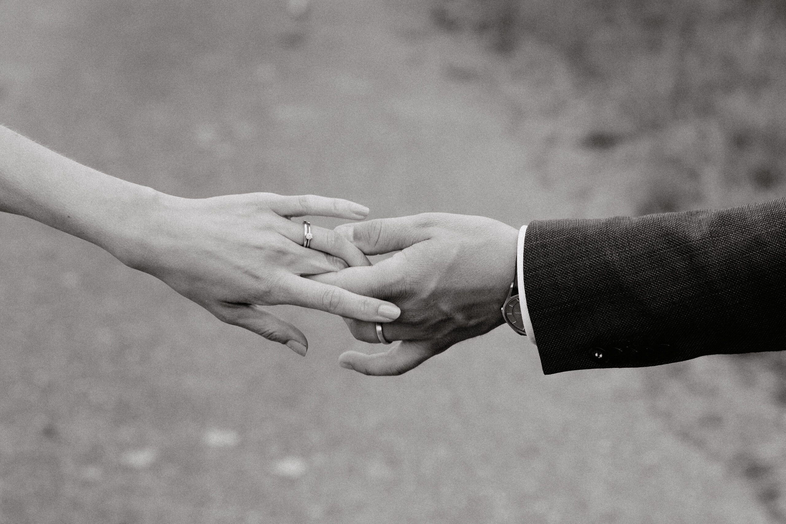 Detailaufnahme der Hände des Brautpaares mit den Eheringen und die schön gemachten Nägel der Braut sind auch zu sehen. Bild in Schwarz weiß. Das Brautpaar spielt mit seinen Händen und hält sich dabei fest. Der Ehebund wird dabei geschürt.