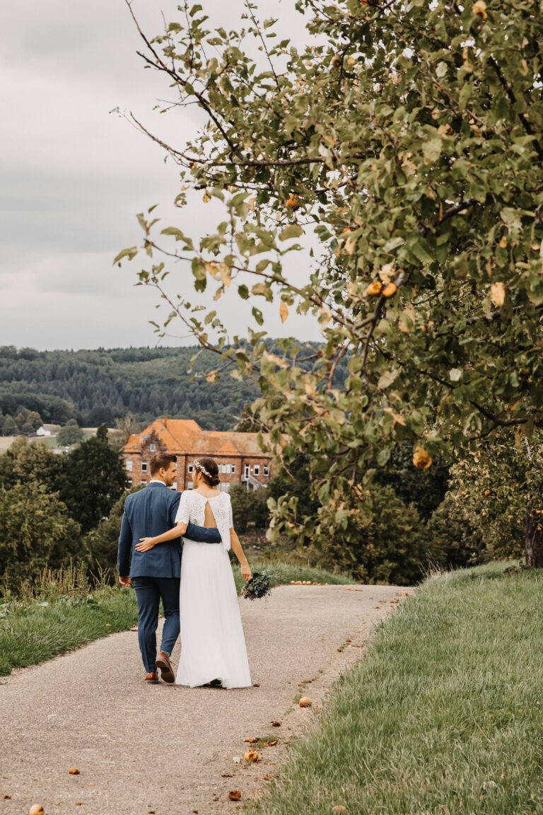 Das Brautpaar läuft arm in Arm und in tiefer Liebe einen Weg entlang. Die Aufnahme fotografiert sie von hinten. Sie schauen sich dabei beide an. Die beiden laufen auf dem Weg in der Nähe von Freiburg umgeben von Feld, grüner wiese und Obstbäumen.