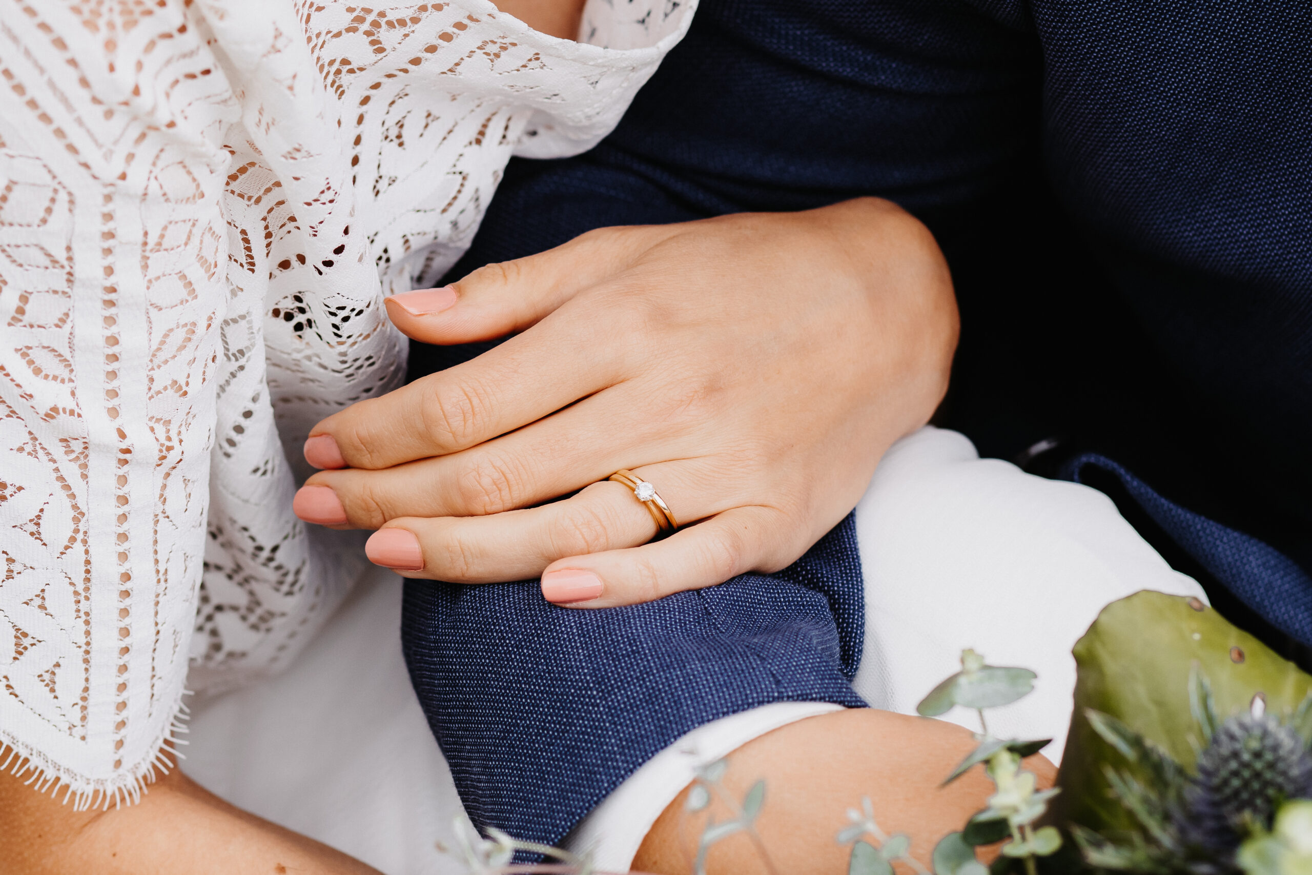 Brautpaar hält sich während dem Brautpaarshooting sitzend die Hand und es ist eine Detailaufnahme der Hände wie die Braut sich bei ihrem Mann einhakt mit der Hand wo auch der Ehering und Verlobungsring zu sehen ist. Der Bräutigam trägt einen blauen Anzug und sie ein weißes Hochzeitskleid.