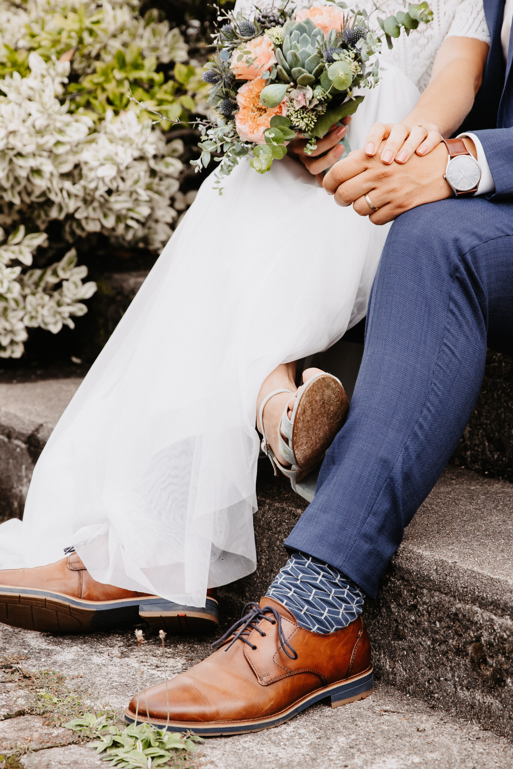 Brautpaar hält sich während dem Brautpaarshooting sitzend die Hand und es ist eine Detailaufnahme des unteren Körper Bereichs. Füsse, Hände und Beine. Der Bräutigam trägt einen blauen Anzug und sie ein weißes Hochzeitskleid.