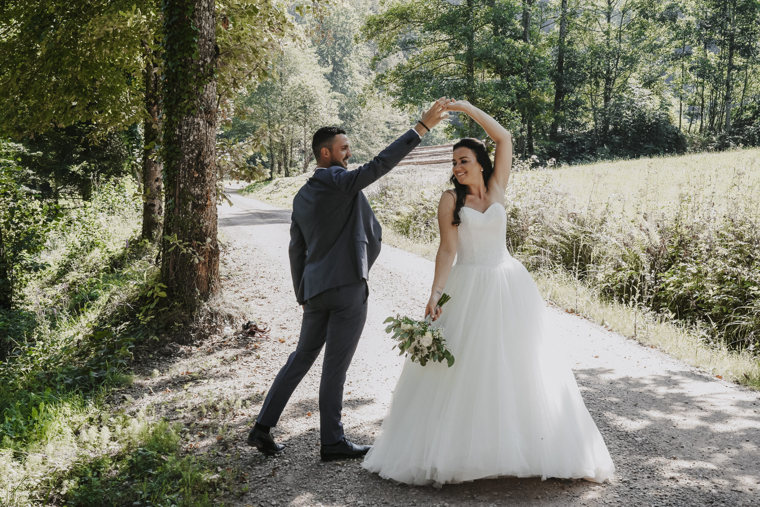 Frau und Mann im Hochzeitsoutfit am Tanzen auf einem Feldweg umgeben von Bäumen.