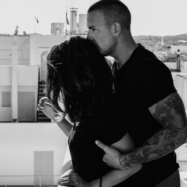Coupleshooting in Ibiza auf einer Dachterasse über den Dächern von San Antoni. Ein Mann umarmt seine Frau von hinten und hält sie fest und kuschelt sich an sie ran. Das Bild ist in Schwarz weiß aufgenommen und sie tragen beide dunkle Kleidung