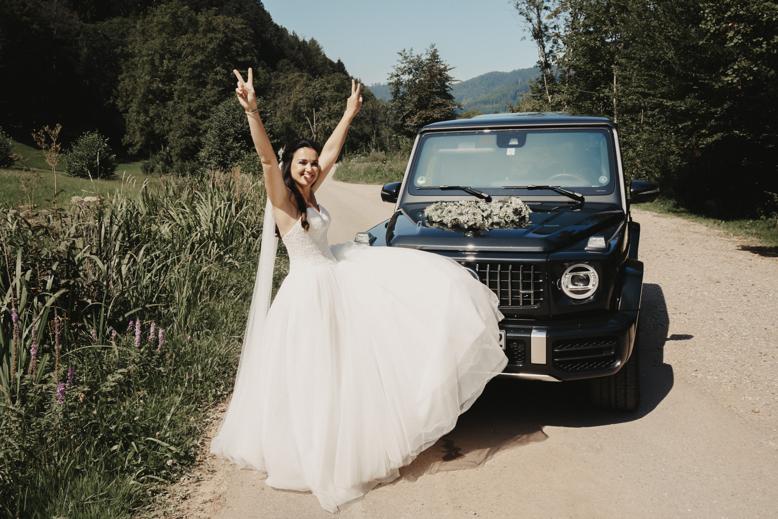 Die Braut post in ihrem großen Brautkleid vor dem Hochzeitsauto. Das Auto ist ein schwarzer Mercedes der in der Natur des Schwarzwalds steht.