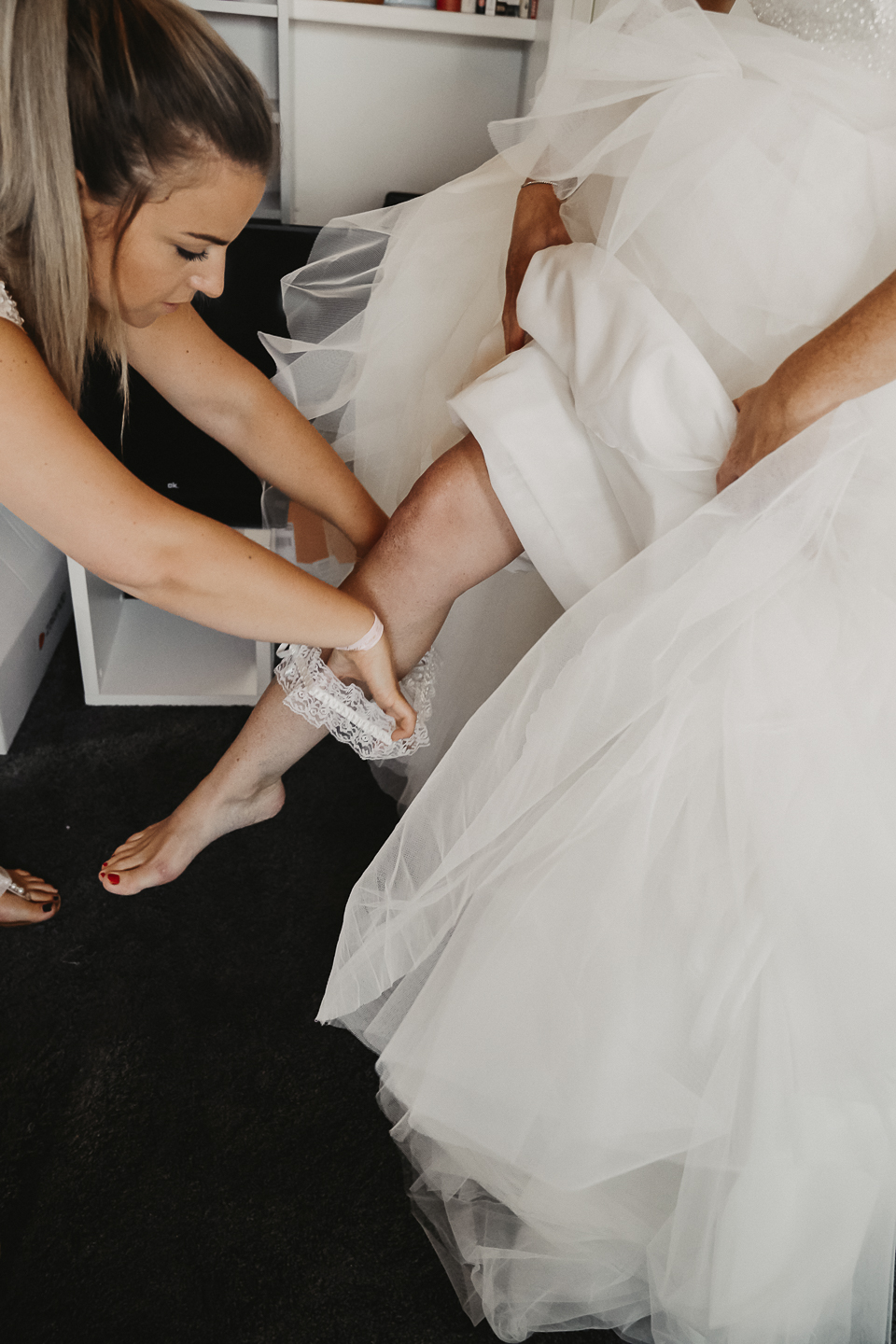 Die Braut wird von ihrer Trauzeugin gerichtet und das Hochzeitskleid und Hochzeitsaccessoires angezogen in ihrem zu Hause. Die Corsage wird festgezogen und das Brautband übergezogen.