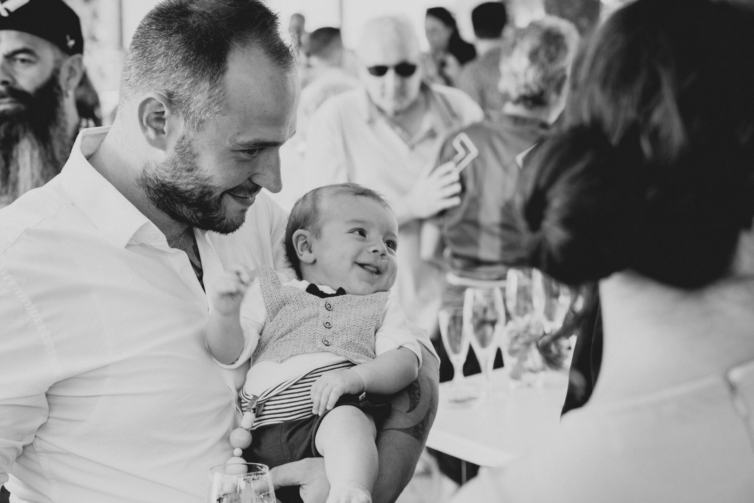 Hochzeitsgast hat seinen kleinen Sohn auf dem Arm der zu anderen Hochzeitsgästen grinst. Sehr intimes und zuckersüsses Foto.
