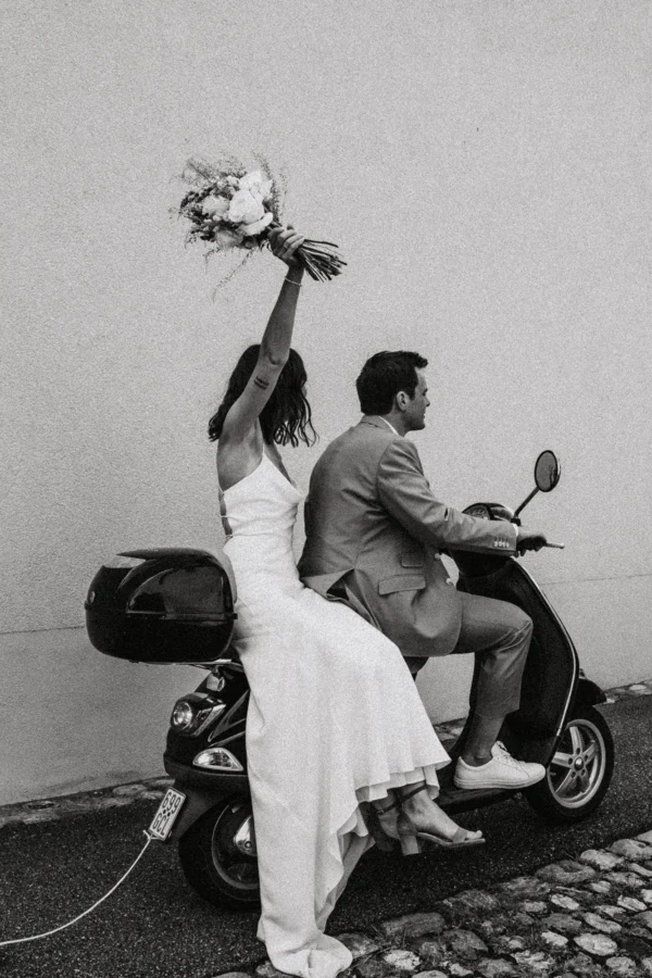 Hochzeutspaar fährt auf ihrer Vespa davon mit klappernden Dosen am Auspuff - Die Braut hält den Brautstrauss in die Höhe